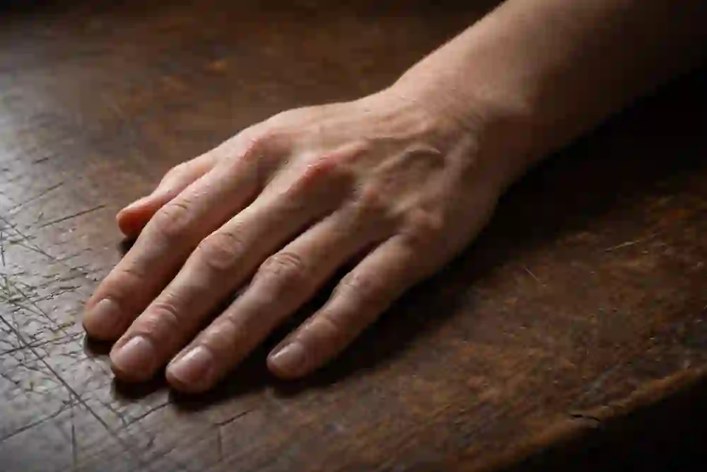 A high-detail candid photo of a woman’s hand resting on a textured wooden table, showing skin wrinkles and natural fingernail texture.