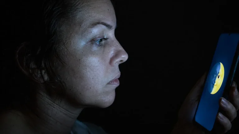 A raw, unedited photo showing a woman’s face in low light, her skin showing visible pores and grit, as she stares at a glowing phone screen featuring the waxing crescent moon emoji, illustrating a digital energy leak.