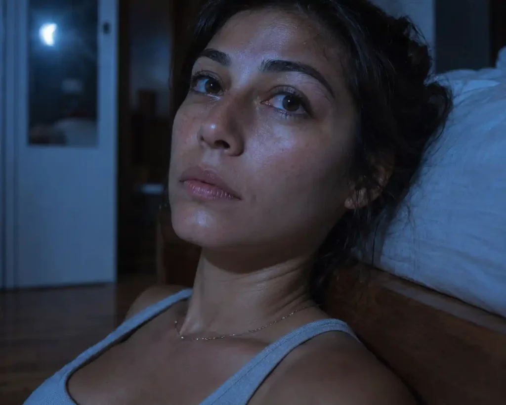 Candid shot of a woman with messy hair sitting by a window in blue moonlight, experiencing red moon cycle spiritual signs.
